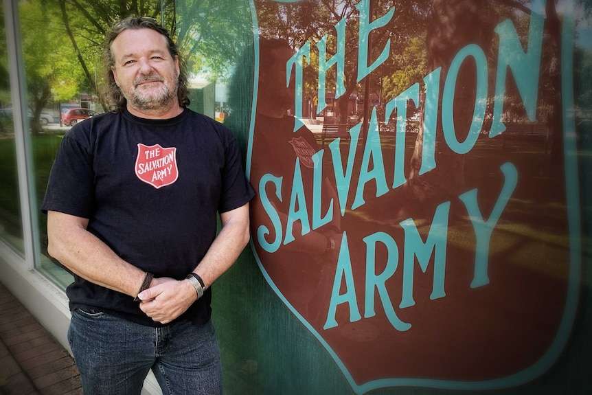 A man with a salvation army shirt leans against a wall.