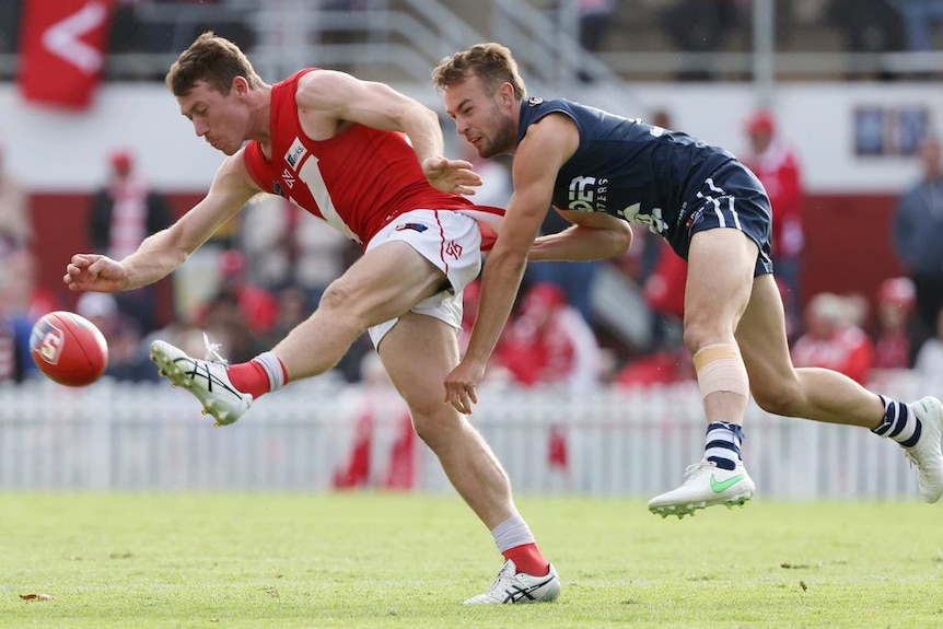 A football player kicks a ball while being tackled by another.