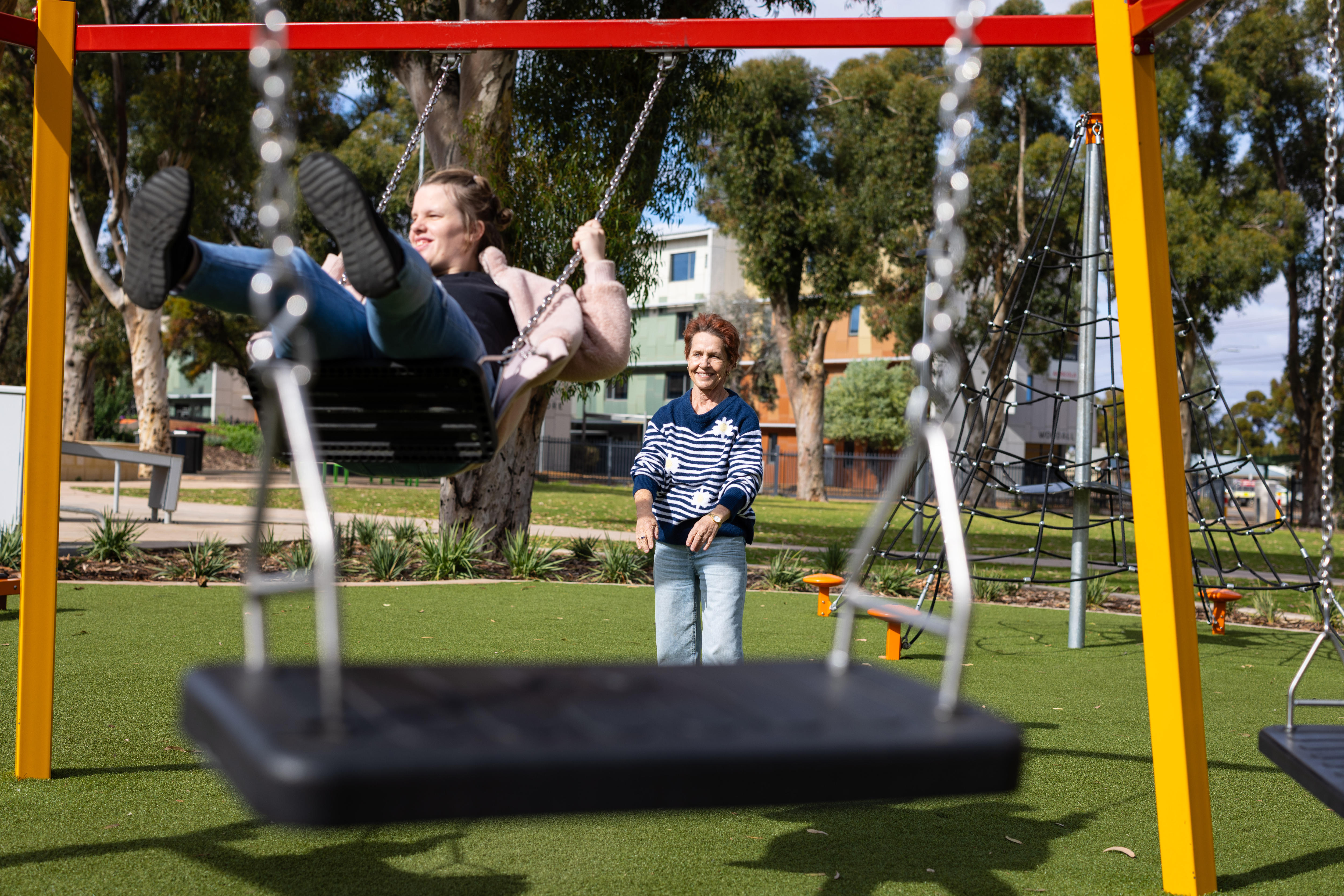An older woman pushes her adult daughter on a swing in a park
