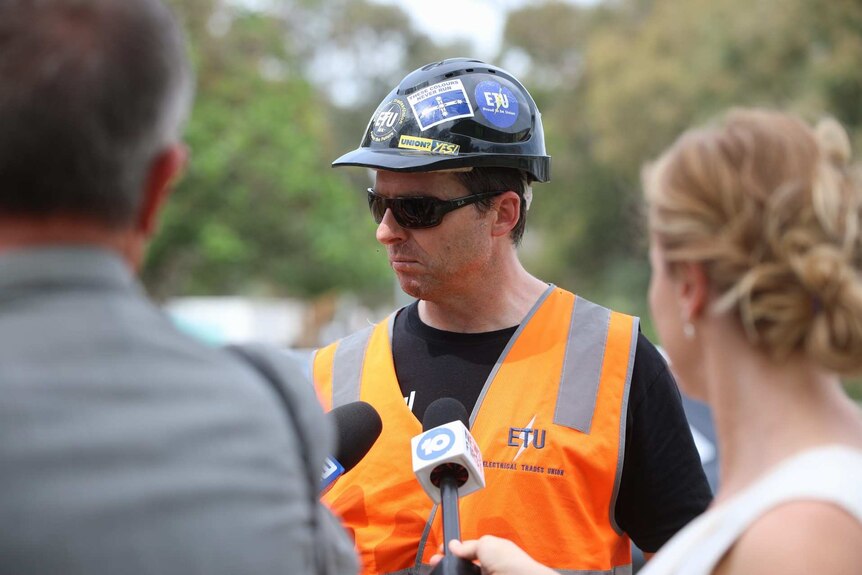 A head and shoulders shot of a man in an orange hi-vis vest and black hard hat talking to reporters.