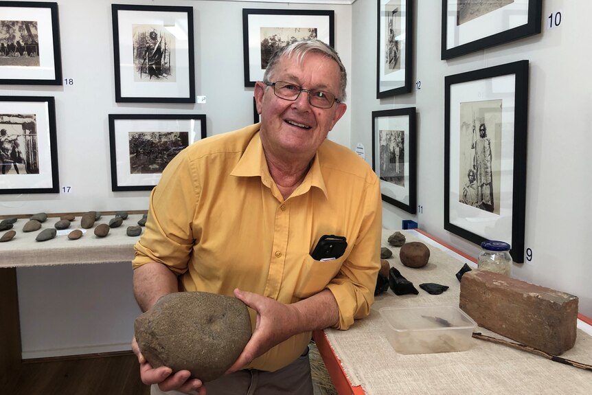 A man in a yellow shirt holds a grinding stone while smiling in a museum 