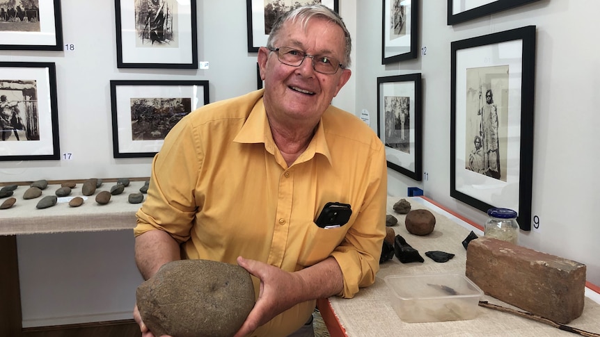 A smiling, bespectacled older man with neat grey hair and a brightly coloured shirt, stands in a room. He holds a large rock.
