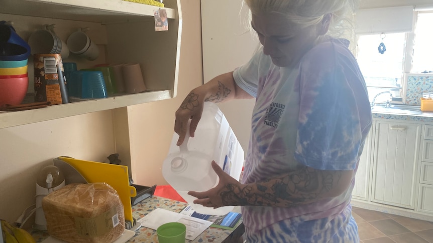 A blonde woman in a multicoloured shirt pours water from a large bottle into a cup, which sits on her kitchen bench.