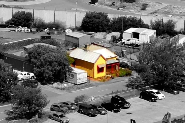 An aerial shot of a yellow and red weathered cottage sandwiched between port infrastructure at Fremantle Port
