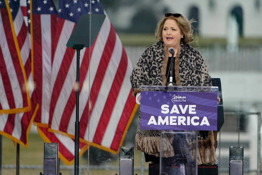 A blonde haired woman is standing and speaking at a podium that has the words 'Save America' on it.