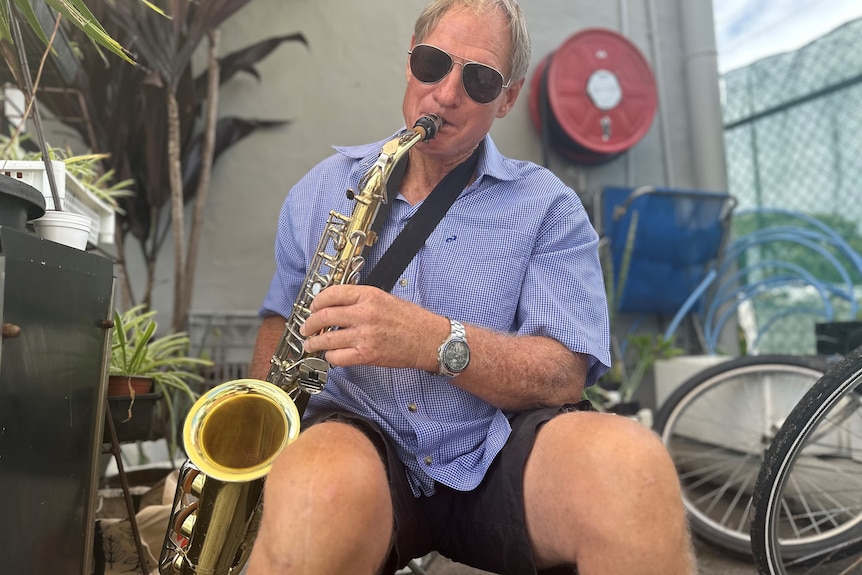 A man sits on a chair in an outdoor courtyard space, playing the saxophone.