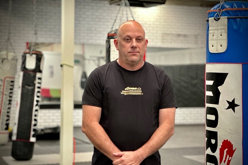 Bald man in active wear t-shirt standing in front of punching bags with serious facial expression