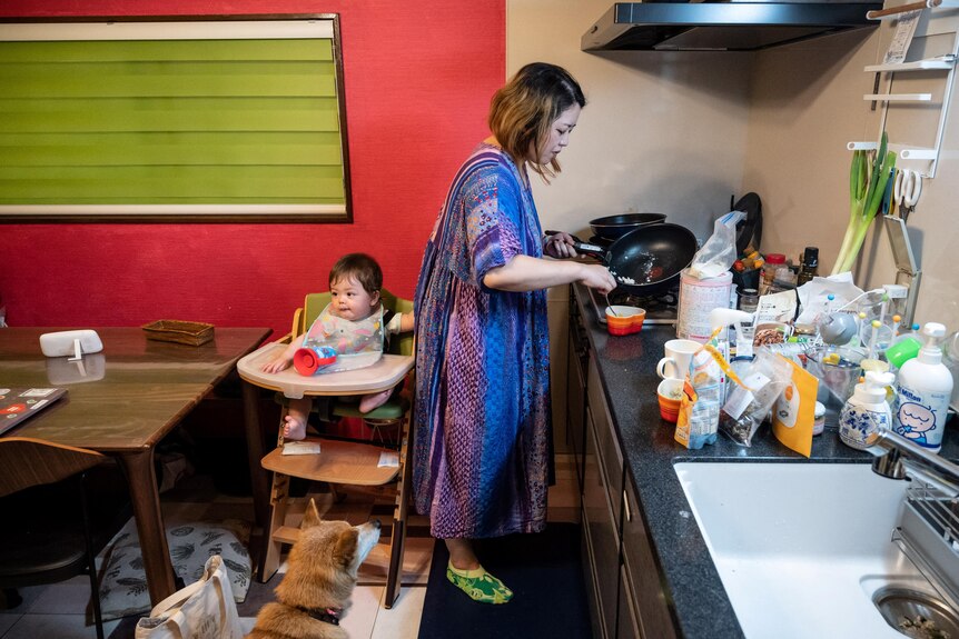 An Asian woman in a vibrant dress stands at the kitchen while behind her a toddler sits in a highchair