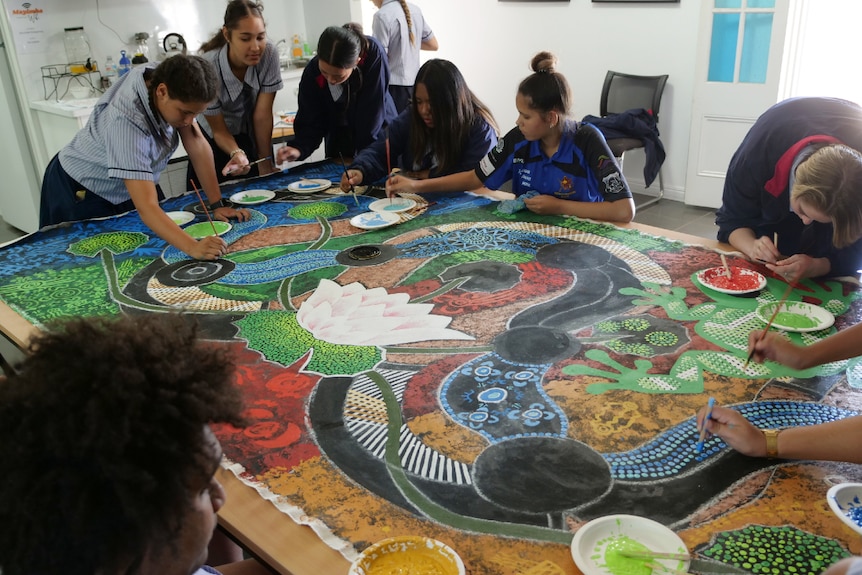 Students leaning over a canvas on a table, painting a colourful artwork.