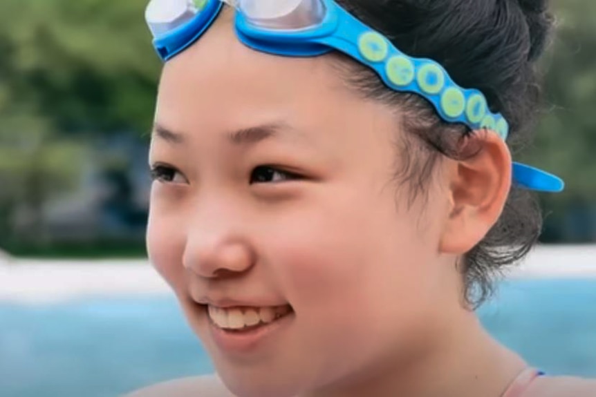 A young Asian girl wearing goggles pulled up to her forehead smiles in a pool