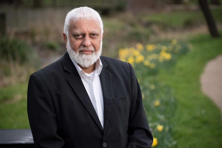 An older man looks directly into the camera who has light brown skin, white hair and a beard, and is dressed in a black suit.