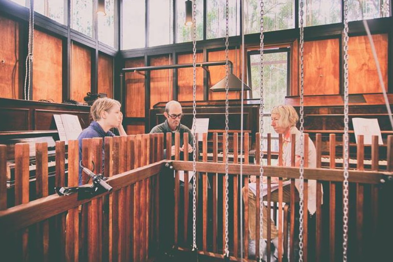 The people face a central void while seated at pianos inside the Piano Mill.