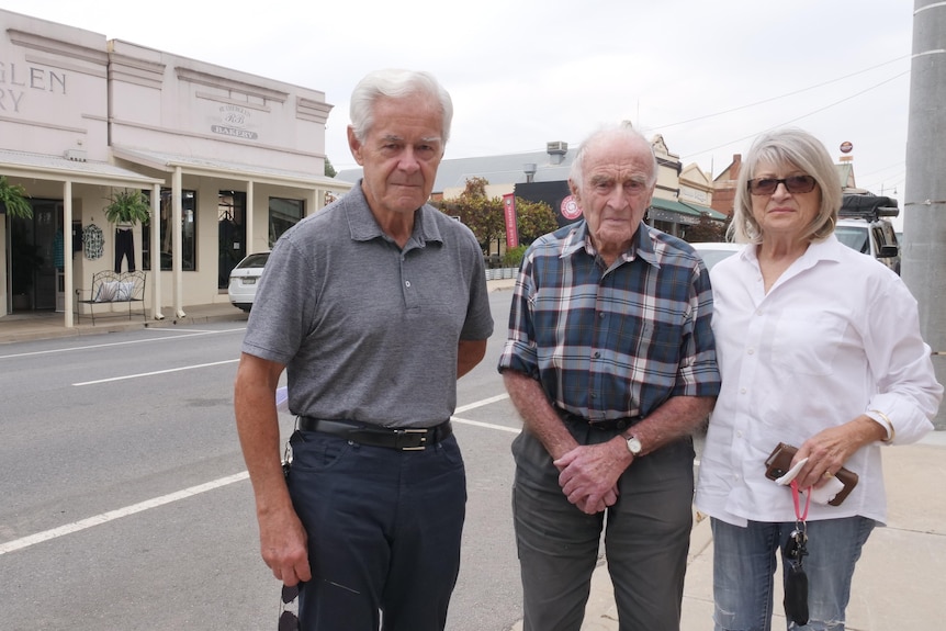 Three older people stand at the edge of the main drag of a country town, all looking solemn.