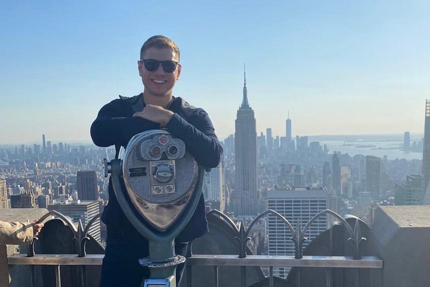 Yair Netanyahu leans on a binocular machine on top of a building with views of NYC skyline including Empire State building