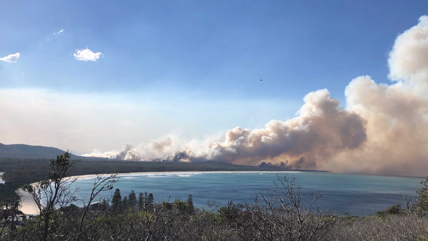Large plumes of smoke rise in the distance of a coastal landscape, with bushland, beaches and water in the foreground.