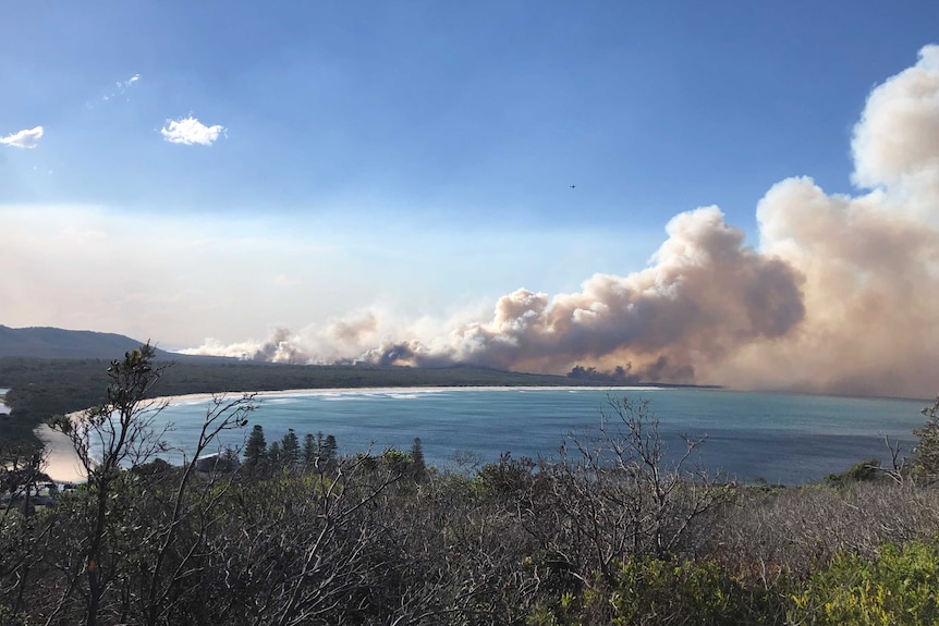 A bay surrounded by bushland with large plumes of smoke billowing in the distance against a blue sky.