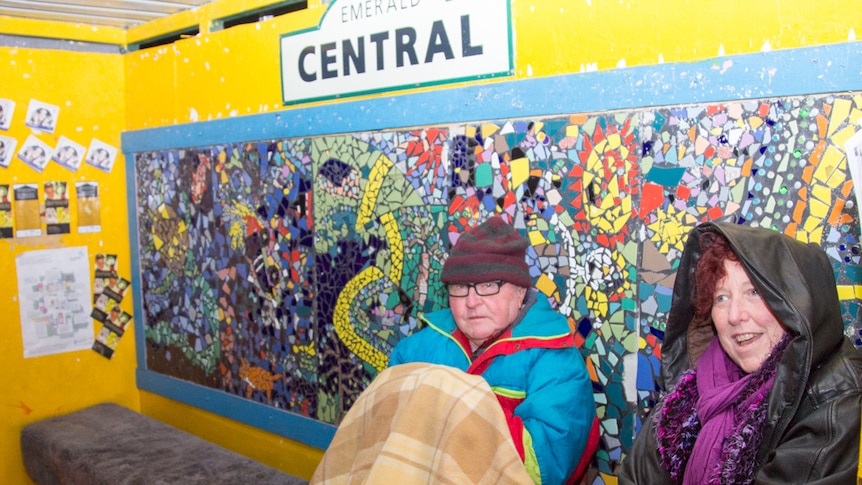 Two people sit in warm clothing under a blanket at a colourful bus shelter.