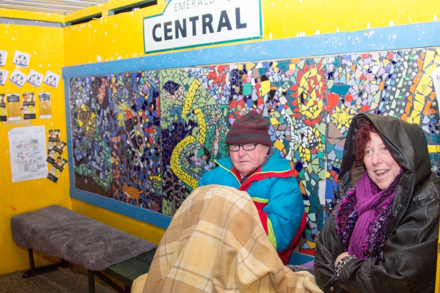 Two people sit in warm clothing under a blanket at a colourful bus shelter.