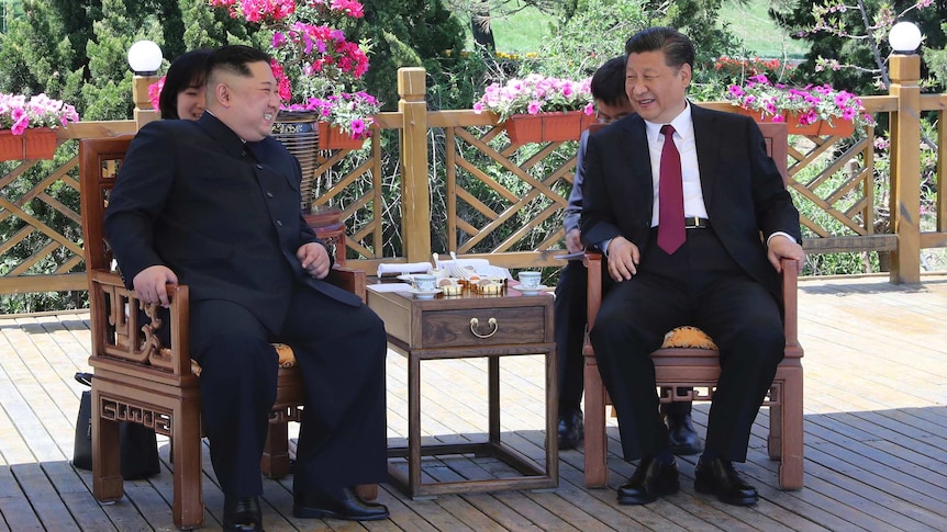 Kim Jong-un and Xi Jinping sit on chairs smiling and drinking tea, with a garden in the background.