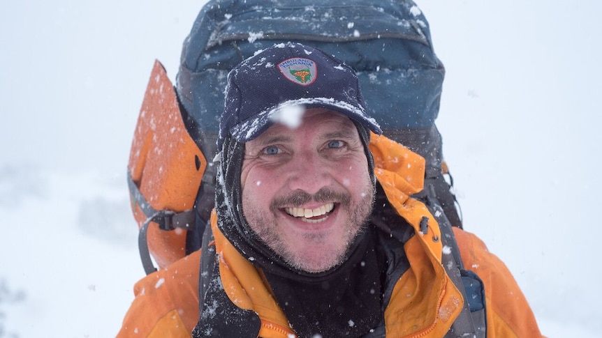 Dave Brown, wearing a bright yellow jacket, a dark cap and backpack, smiles at the camera as snow falls around him.