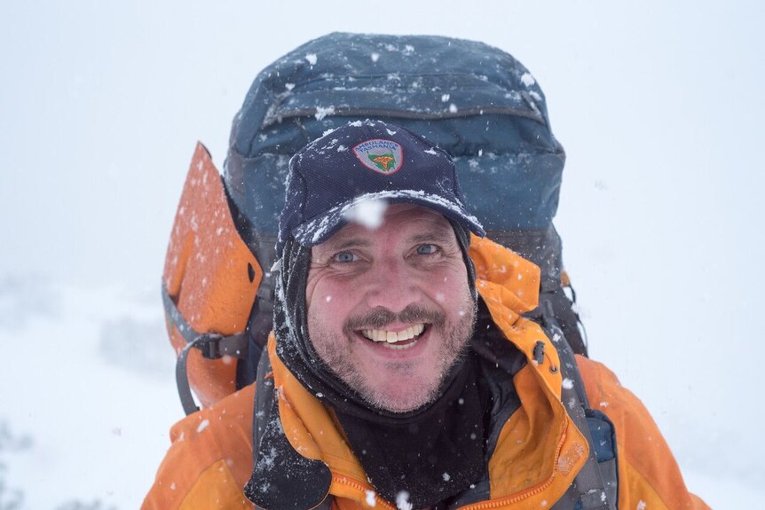 Dave Brown, wearing a bright yellow jacket, a dark cap and backpack, smiles at the camera as snow falls around him.