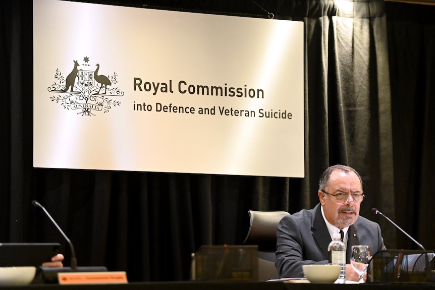 a man wearing glasses speaking into a microphone sitting behind a desk