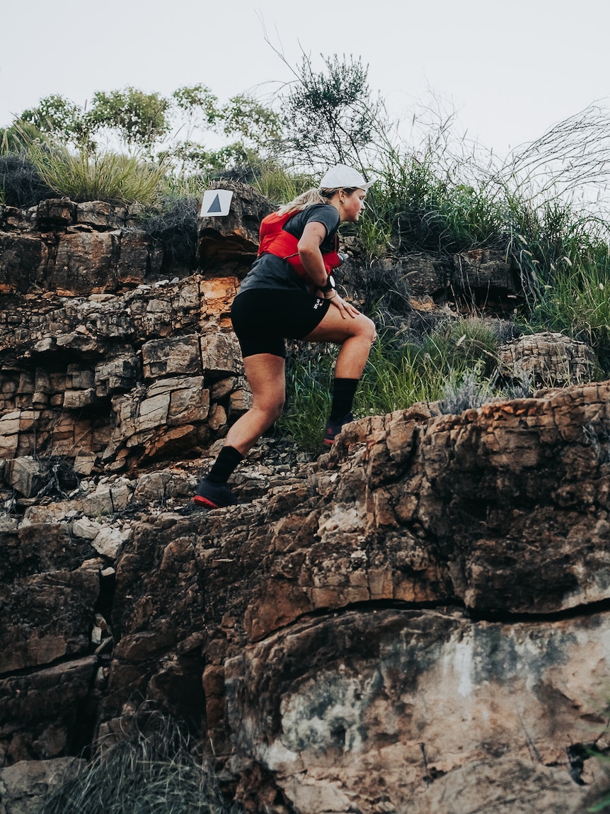 Woman wearing black with red running pack and a white cap traversing rocks