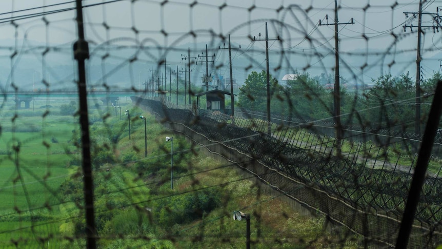 A large fence topped with barbed wire extends along green land towards mountains.