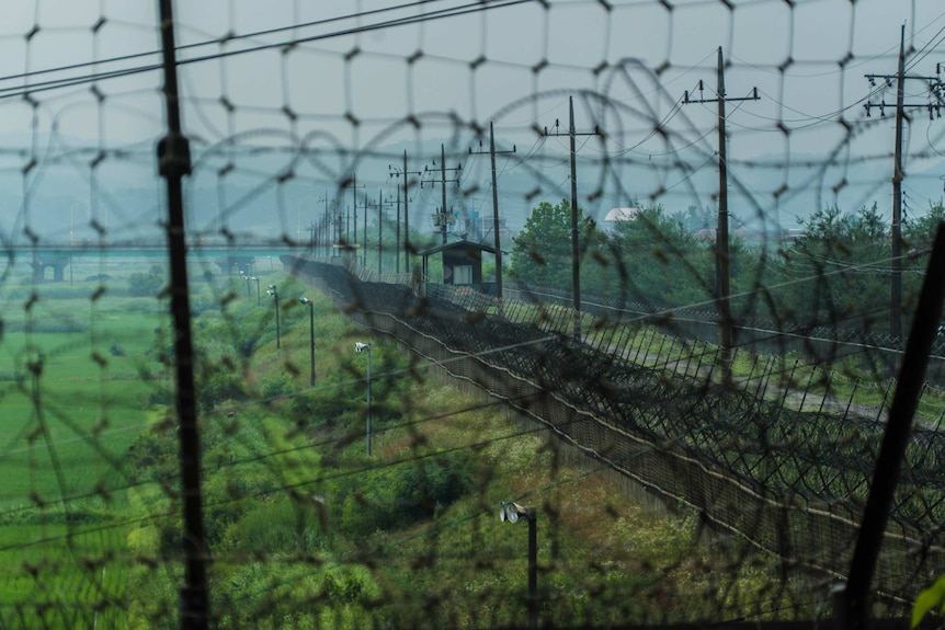 A large fence topped with barbed wire extends along green land towards mountains.