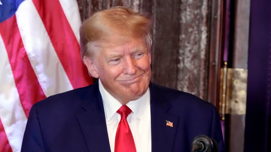 A tanned older man with yellow hair smirks in front of an American flag.