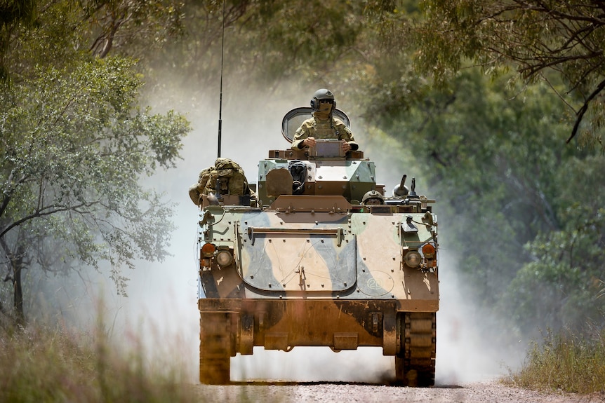 An Australian Army armoured personnel carrier drives towards the camera, kicking up dust, helmeted crewman standing in turret