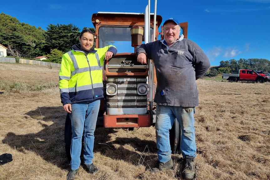 Young woman dressed in high vis and older man lean on the front of a retro tractor in a dead field