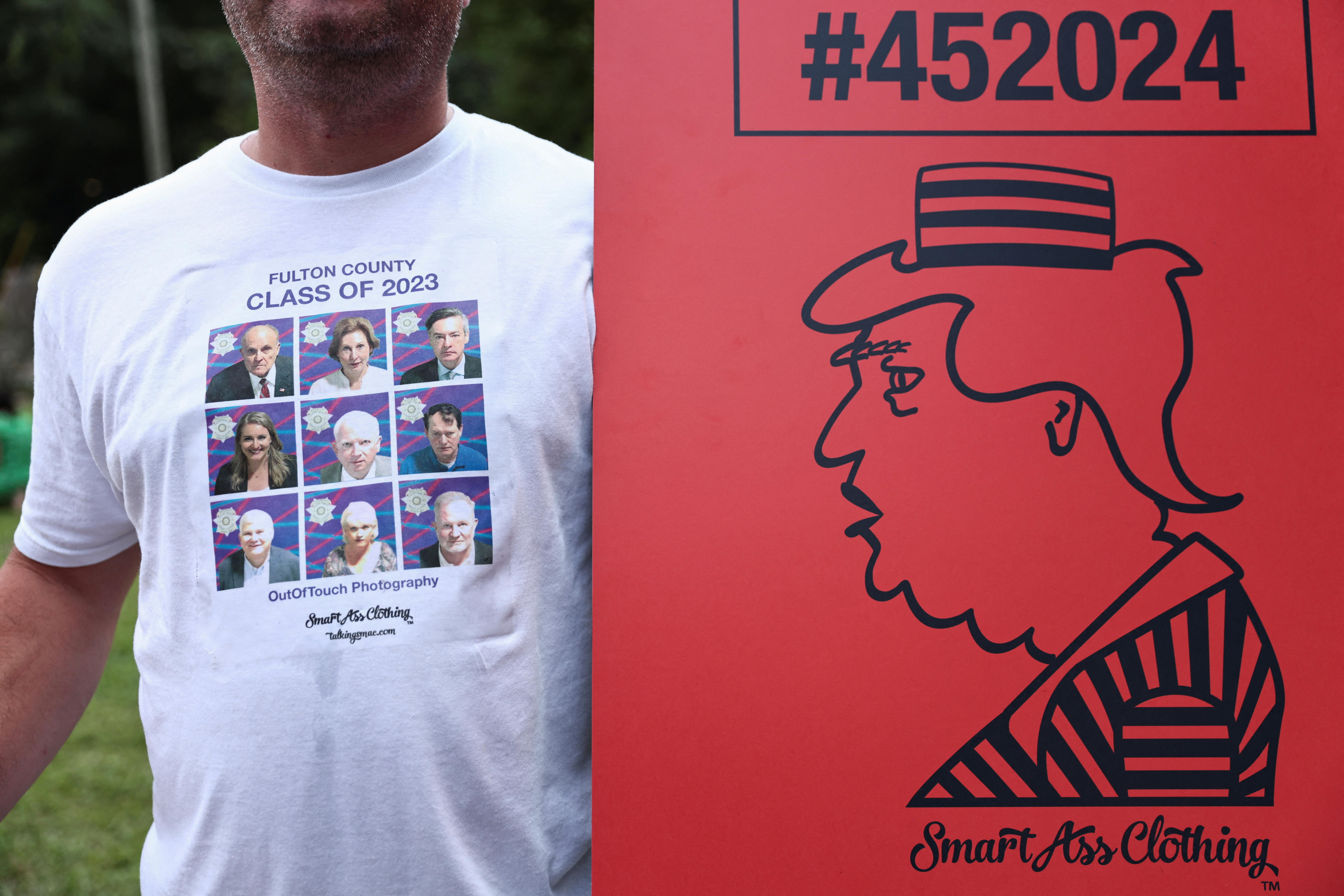 A man wearing a T-shirt with the mugshots of Donald Trump's alleged co-conspirators and a poster with a drawing of him in prison