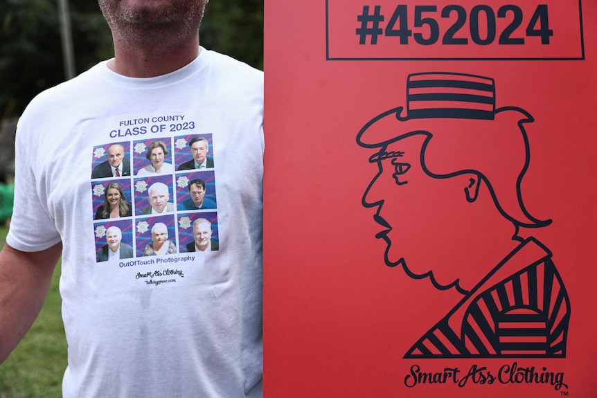 A man wearing a T-shirt with the mugshots of Donald Trump's alleged co-conspirators and a poster with a drawing of him in prison