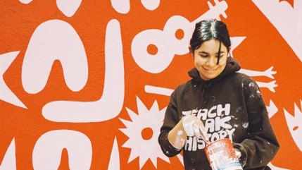 A woman with dark hair and paint-covered clothes stands in front of a mural smiling.