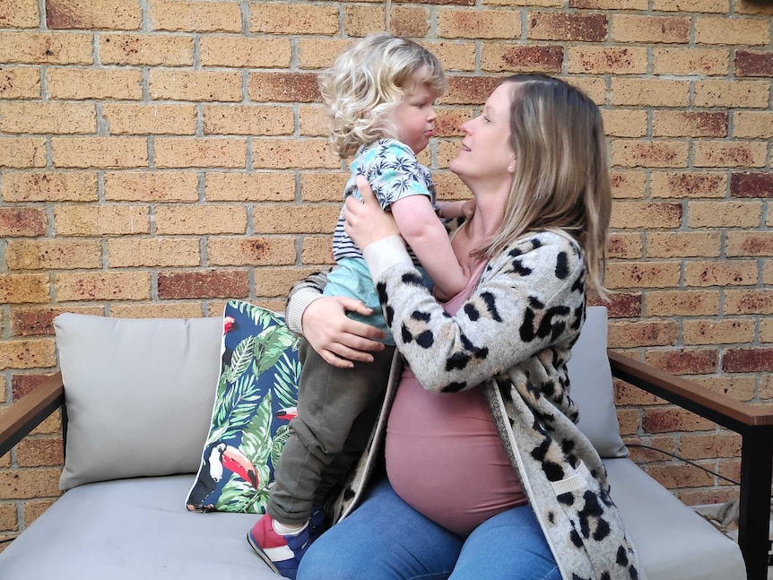 A pregnant woman sitting on a bench hugs her young son who is standing next to her.