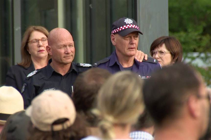 A Victoria Police officers with his arm around a CFA member standing in front of a large crowd.
