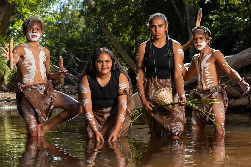 Four young people, two boys and two girls, stand knee deep in water dressed in traditional paint.