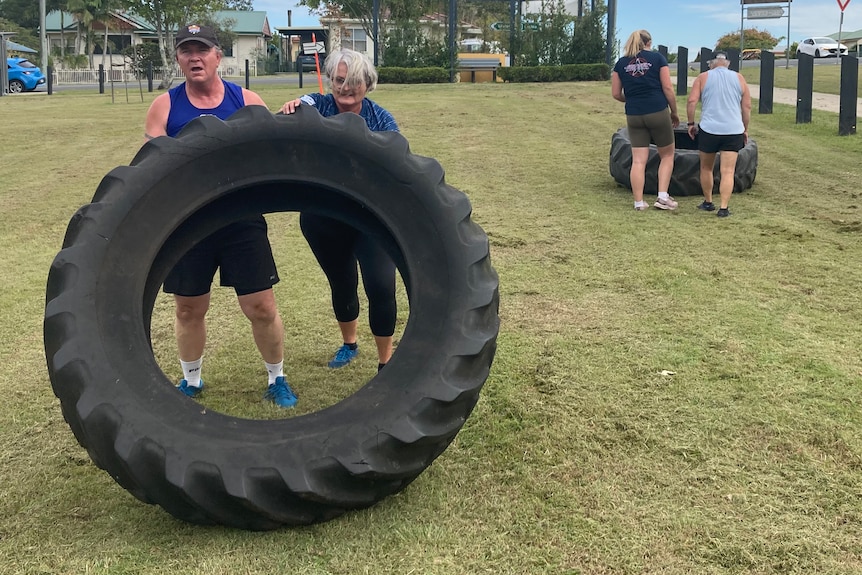 Two people push a very large tyre upright toward the camera, while behind them two people push another tyre the other way.