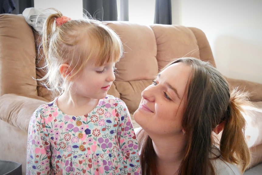A dark haired woman in a living room smiles at her young, fair-haired daughter.