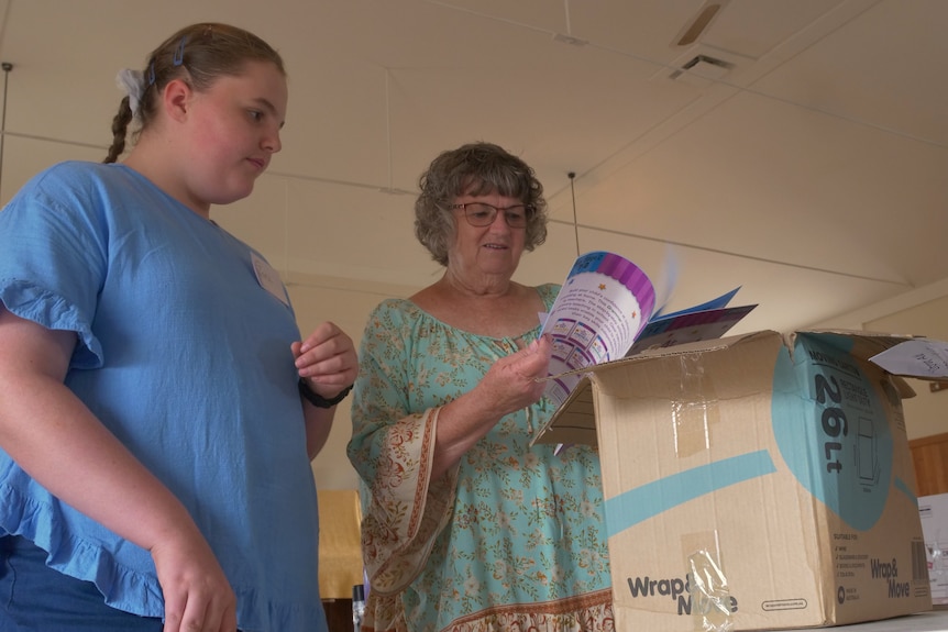 Two woman wearing blue shirts both look into a box of books, grinning.