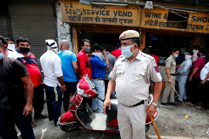 A police officer wearing a mask and carrying a bamboo stick stands in the middle of a crowd outside a wine shop