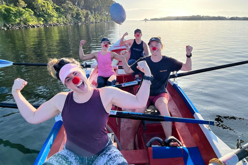 Women wearing red noses flex their muscles in a row boat on water