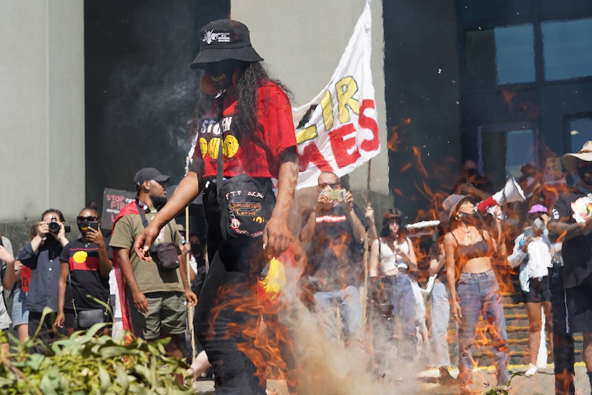 A protester wearing a dark mask stands in front of burning leaves as demonstraters stand in the background