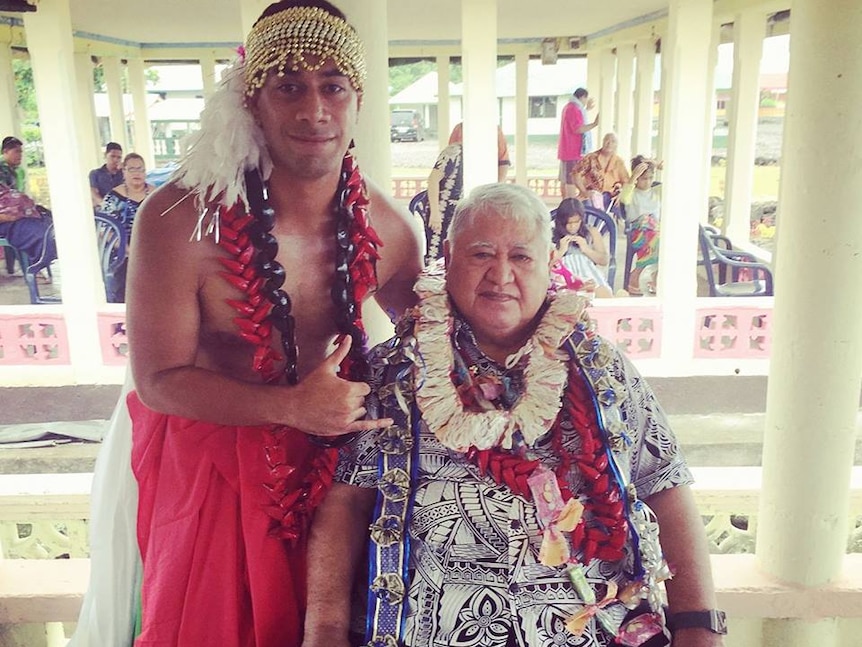 A shirtless man wearing traditional head-dress, sarong and necklaces poses in an open Samoan house with another seated man.