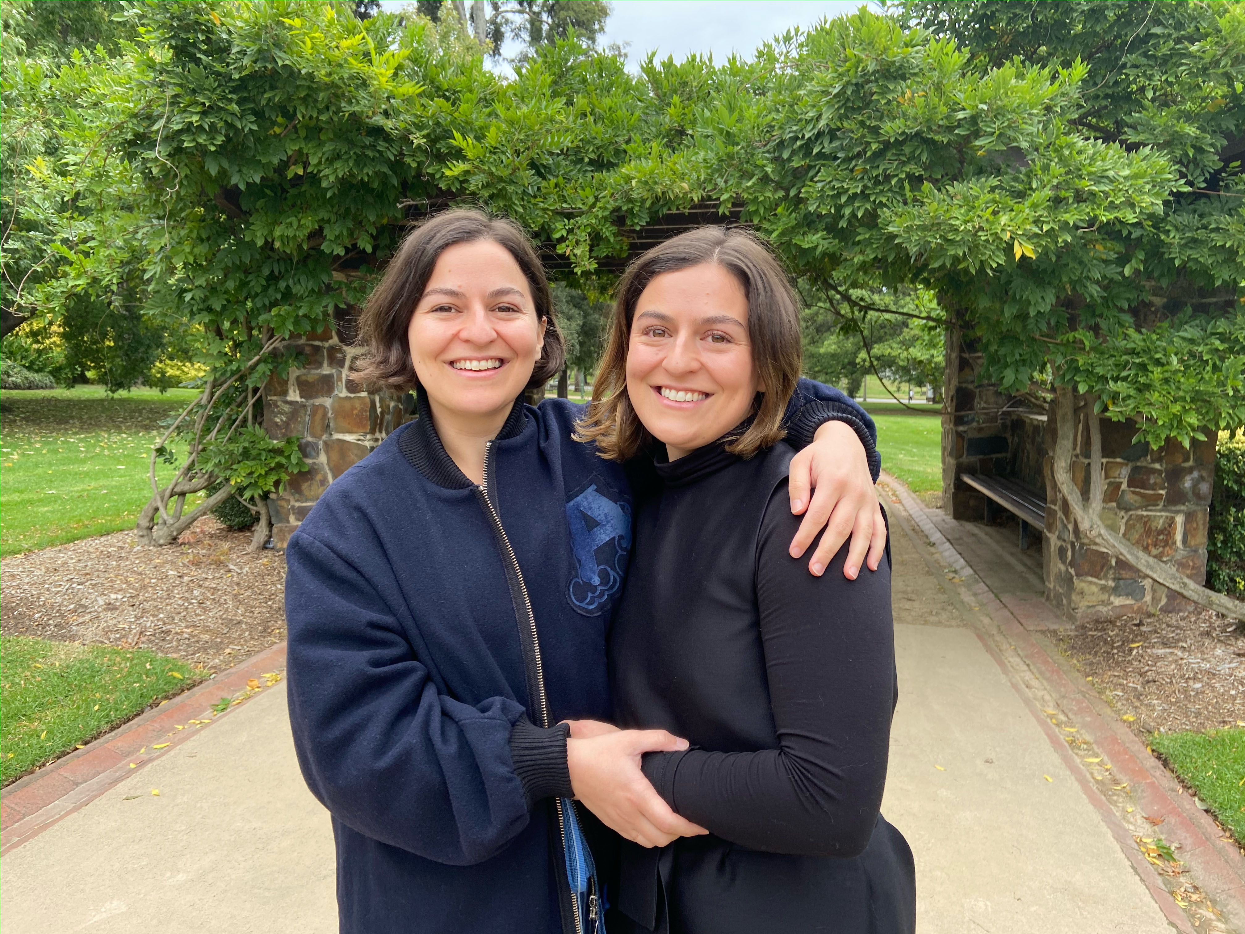 Twin adult sisters stand in a garden with their arms around each other.