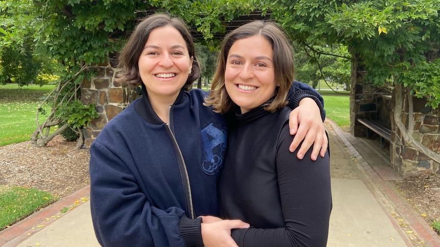 Twin adult sisters stand in a garden with their arms around each other.