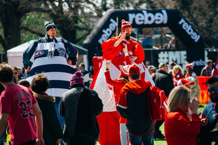 Two men in large inflated Geelong and Sydney AFL team suits walk among fans in morning sunlight.