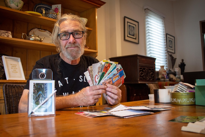An older man with grey hair, beard and black glasses holds an array of postcards