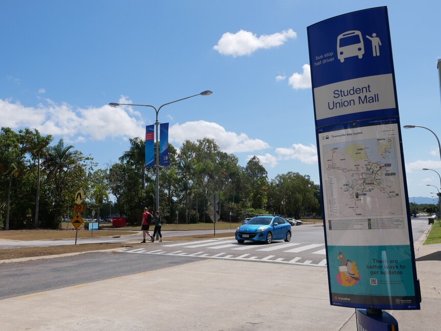 A tall narrow bus sign by a cross walk on a four lane road during the day. 